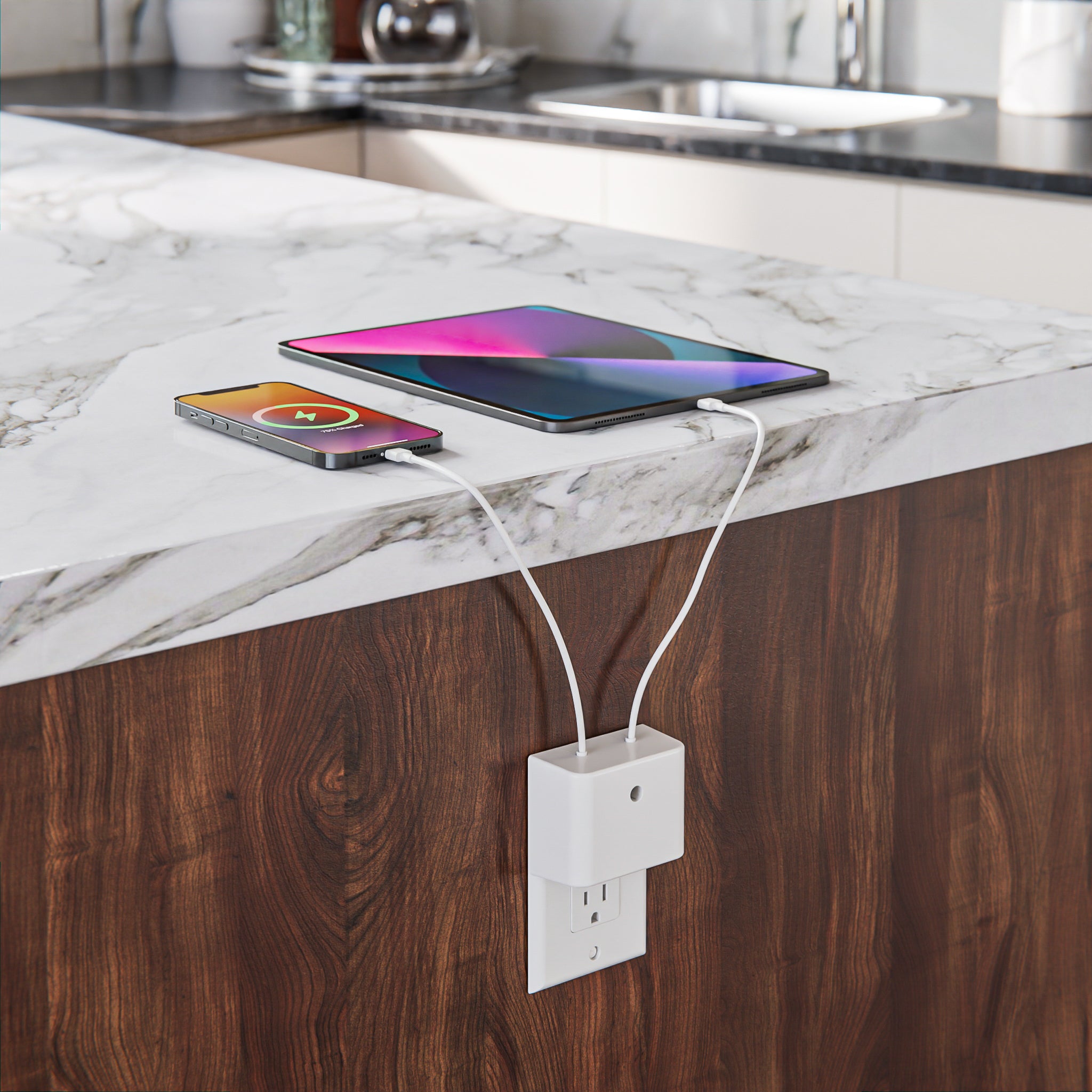 This image showcases the Permaplug Charger Lock Decora model installed on a decorator-style outlet in a kitchen setting. The outlet is positioned beneath a marble countertop, with a tablet and smartphone charging on the counter. The Decora model is distinguished by its top and bottom screw holes, which secure it to the decorator-style outlet. The cables are neatly connected, and the devices are actively charging, highlighting the product's compatibility and efficiency in modern spaces.