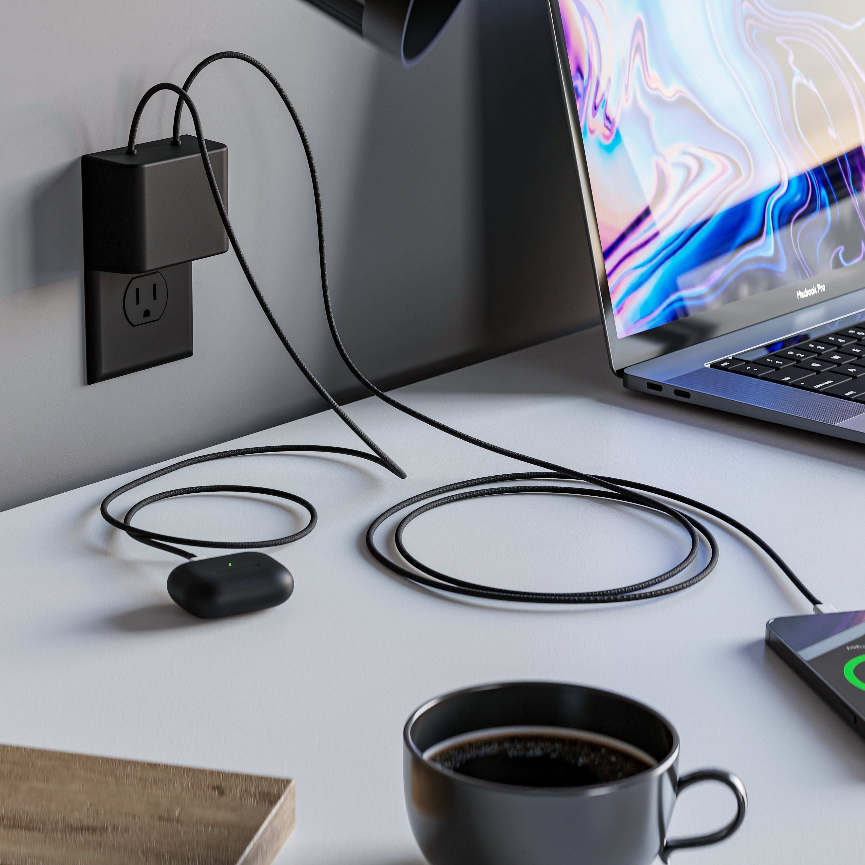 This image features the limited edition black Permaplug Charger Lock Standard model, installed on a standard black wall outlet. The setup includes a MacBook Pro and wireless earbuds on a modern desk, both charging with black cables connected to the charger lock. The central screw hole on the charger lock indicates its compatibility with standard outlets. The all-black charger lock, cables, and outlet create a sleek and minimalist aesthetic, blending seamlessly with the modern workspace. A cup of coffee and