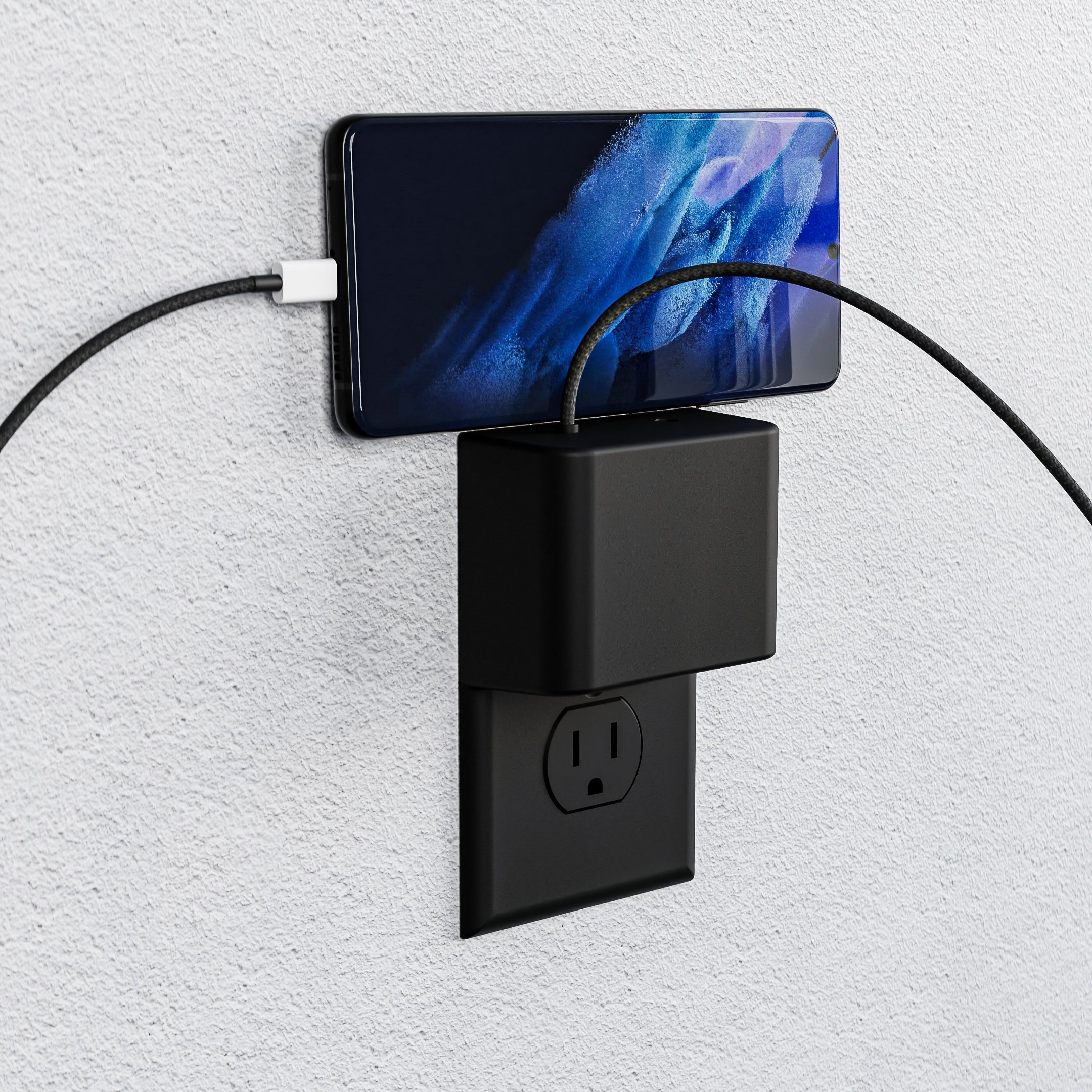 This image showcases the limited edition black Permaplug Charger Lock Standard model, installed on a standard outlet against a textured wall. A smartphone is securely positioned on top of the charger lock while charging, with two cables attached. The black-on-black setup creates a sleek, seamless look, emphasizing both style and functionality while ensuring secure, organized charging in a modern setting.